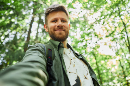 Smiling man taking a selfie in a lush green forest surrounded by tall trees. Appearing relaxed while enjoying nature with sunlight filtering through leavesの写真素材