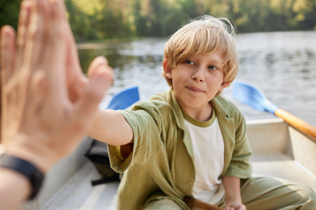 A young boy with blond hair is sitting in a canoe on a lake, giving a high-five to someone. The background features peaceful scenery with trees and waterの写真素材