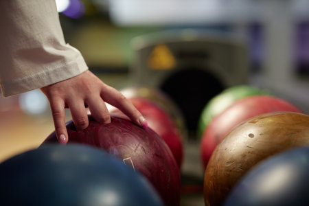 Hand reaching for vibrant bowling ball in colorful, well-lit bowling alley with various balls and blurred background creating dynamic atmosphereの写真素材