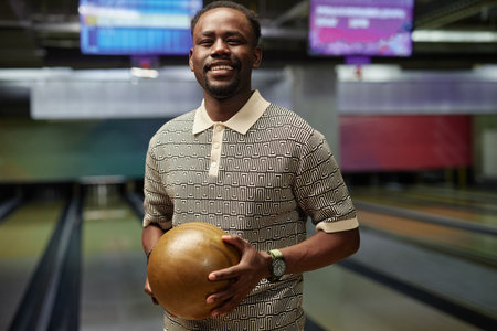African American man holding bowling ball while standing at bowling alley, smiling warmly, wearing patterned shirt and wristwatch, bowling lanes visible in backgroundの写真素材