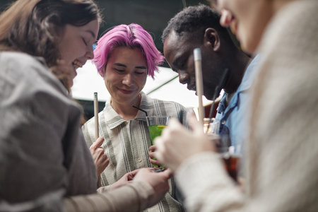 Group of young adults enjoying time together at a modern gathering event while chatting and using gadgets. Multiracial friends forming close connectionsの写真素材