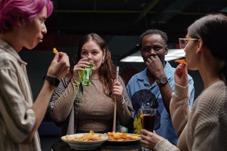 Group of friends enjoying snacks and drinks in an indoor setting while engaging in conversation and using variety of gadgets. Multi-ethnic individuals experiencing a relaxed momentの写真素材