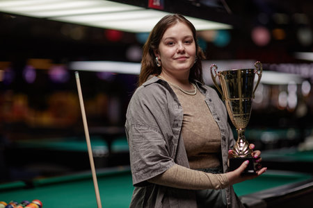 Portrait of smiling woman holding trophy in pool hall, surrounded by pool tables and colorful...