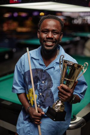 African American man smiling and holding trophy while leaning on pool cue by pool table indoors with...