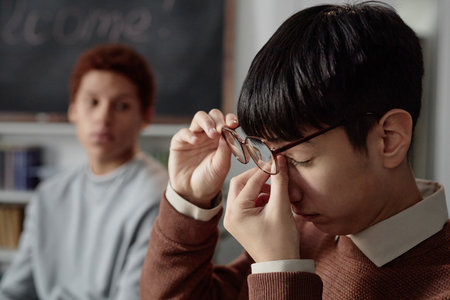 Close up portrait of tired teenage boy suffering from headache in school classroom and taking off...