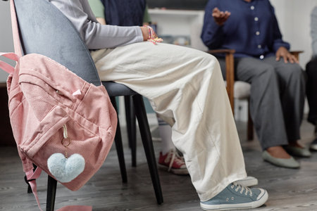 Side view low section shot of teenager sitting on chair during support group meeting with focus on canvas shoes and wide leg pants copy spaceの写真素材