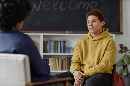 Portrait of rebellious teenage boy talking to therapist or school counsellor while sitting on chair and smiling during one on one meeting copy spaceの写真素材