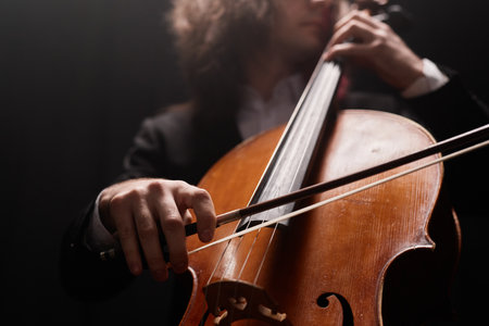 Cropped shot of unrecognizable talented male cello player performing classical music in dark recording studio with dim light, focus on violoncello bowの写真素材