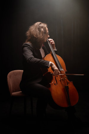 Vertical full shot of male cello player in black tuxedo performing classical music while sitting in chair in dark studio with top light, copy spaceの写真素材