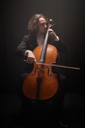 Vertical full shot of professional male musician playing cello solo performing classical music in dark studio with top lightの写真素材
