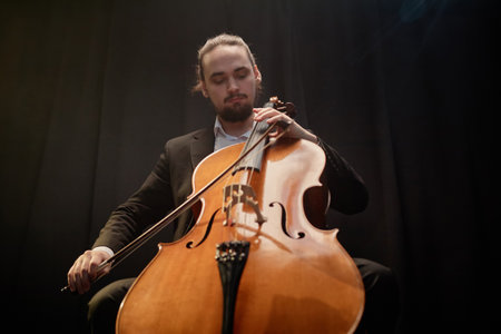 Low angle shot of professional musician playing cello during solo concert against black curtains...