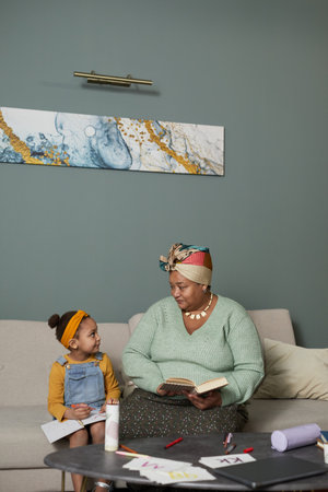 Vertical portrait of African-American grandmother reading book to cute little girl in minimal home...