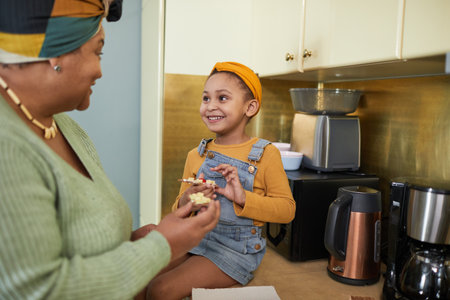 Portrait of cute African-American girl sitting on kitchen counter and enjoying healthy snack with grandmother in cozy home interior, copy spaceの写真素材