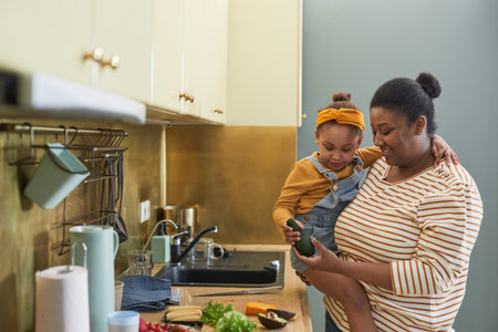 Waist up portrait of happy African-American mother holding daughter while cooking together in kitchen interior, copy spaceの写真素材
