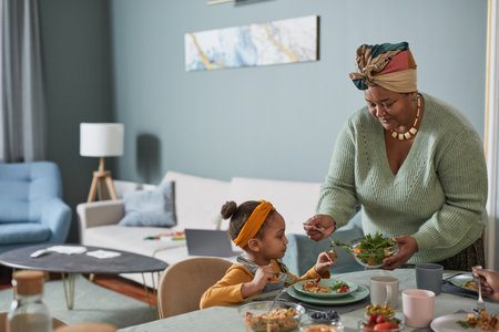 Portrait of African-American family enjoying dinner together in minimal home interior, focus on grandmother serving food to cute little girl, copy spaceの写真素材