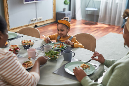 Portrait of cute African-American girl eating food at table and enjoying dinner with family in cozy home interior, copy spaceの写真素材