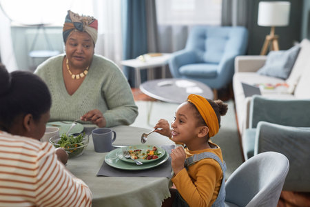 Side view portrait of cute African-American girl eating food at table and smiling happily while...