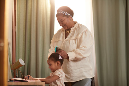 Side view portrait of smiling African-American grandmother combing hair of cute little girl while standing by mirror in cozy home interiorの写真素材