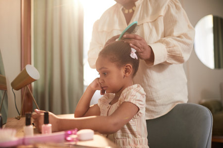 Cropped portrait of African-American grandmother combing hair of cute little girl by mirror in cozy home interiorの写真素材