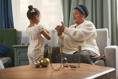 Side view portrait of senior African-American woman showing tricks to cute little girl while playing together in cozy home settingの写真素材