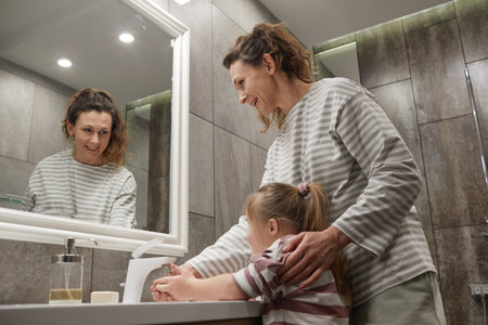 Side view portrait of smiling mother helping little girl with down syndrome wash hands at sink in bathroom, copy spaceの写真素材