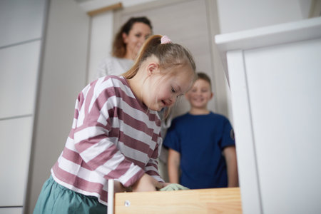 Side view portrait of cute girl with down syndrome opening drawer while enjoying cleaning with brother and mom, copy spaceの写真素材