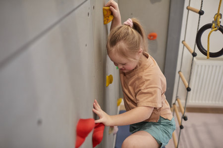 High angle view at cute girl with down syndrome climbing wall while exercising in sports set at home, copy spaceの写真素材