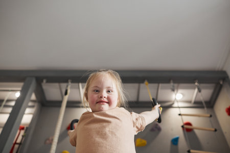 Low angle portrait of cute girl with down syndrome swinging on rings and enjoying exercise at home or at sports center while looking at camera, copy spaceの写真素材