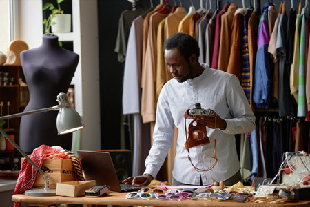 Male African American thrift store assistant in classy shirt using laptop putting up vintage photo...