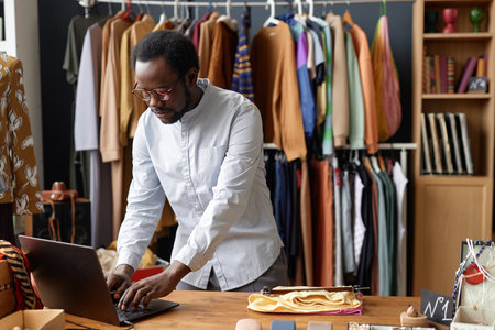 Medium shot of African American salesman using laptop for selling second hand clothes and keeping account in social networks at thrift store checkout, copy spaceの写真素材