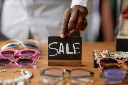Close up on hand of African American salesman setting sale sign on wooden display table cluttered with vintage sunglasses and fashion accessories at second hand shop, copy spaceの写真素材