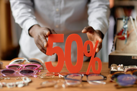 Close up on hands of African American salesman setting fifty percent discount red sign on wooden display table cluttered with vintage sunglasses and fashion accessories at second hand shop, copy spaceの写真素材