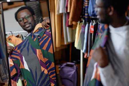 Reflection in mirror of smiling African American man trying vintage summer shirt while shopping at second hand store, copy spaceの写真素材