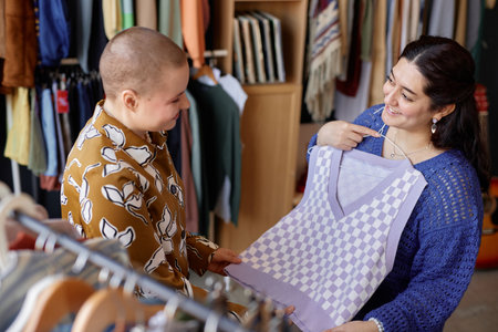 Side view of smiling Middle Eastern woman shopping at thrift store discussing vintage sweater vest with buzz cut female friend, while choosing clothes at second hand shopの写真素材