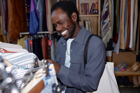Portrait shot of young smiling African American man in gray trendy shirt choosing clothes shopping at second hand store, camera flash, copy spaceの写真素材