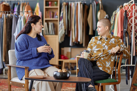 Side view of two young female friends dressed in vintage outfits chatting over cup of coffee discussing fashion while sitting in second hand chairs at thrift store, copy spaceの写真素材