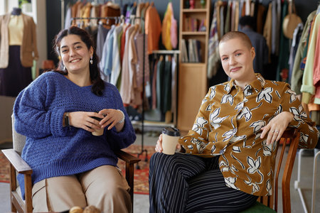 Portrait shot of two Gen Z women dressed in vintage clothes sitting in chairs holding paper coffee...