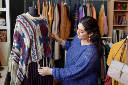 Young Middle Eastern female sales assistant in blue vintage sweater displaying woolen poncho on mannequin at second hand shopの写真素材