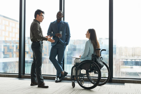 Full length portrait of young businesswoman in wheelchair chatting with male colleagues against window in office, copy spaceの写真素材