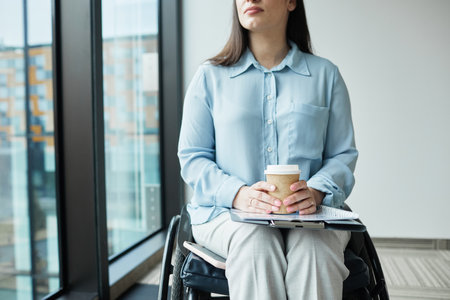 Cropped portrait of pensive businesswoman in wheelchair enjoying coffee by window in office, copy spaceの写真素材