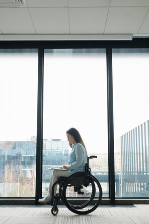 Graphic full length portrait of young businesswoman in wheelchair reading documents by window in...