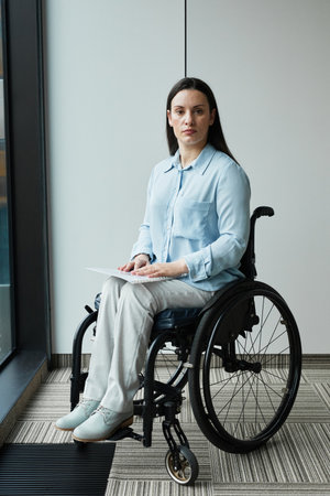 Vertical full length portrait of young businesswoman in wheelchair looking at camera while posing by...