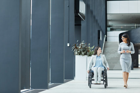 Full length portrait of young businesswoman in wheelchair talking to female colleague while moving towards camera in office lobby, copy spaceの写真素材