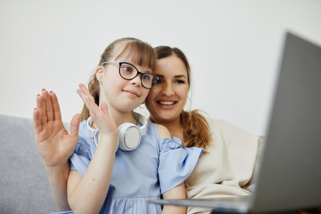Portrait of cute girl with disability waving hello at camera while using video chat with mom, copy spaceの写真素材