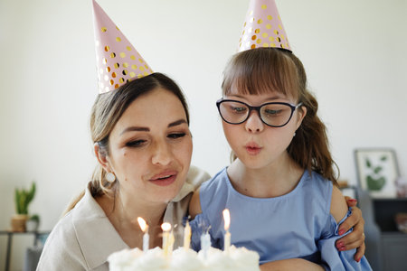 Portrait of happy mother and daughter with disability celebrating birthday online and blowing candles to make wishの写真素材