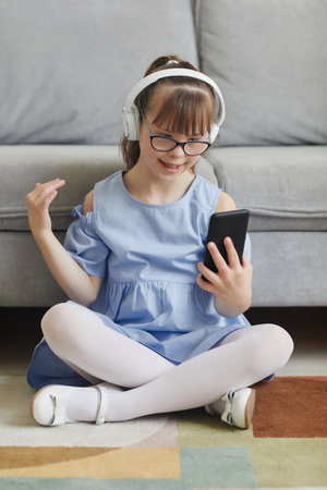 Full length portrait of carefree girl with down syndrome listening to music at home while sitting on floor and using smartphoneの写真素材