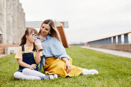 Full length portrait of happy mother and daughter with down syndrome sitting on green lawn outdoors together, copy spaceの写真素材
