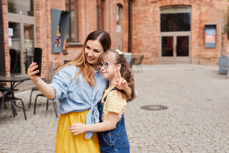 Colorful waist up portrait of happy mother and daughter with down syndrome taking selfie together outdoors in city, copy spaceの写真素材