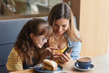 Warm-toned portrait of loving mother and daughter with down syndrome using smartphone while enjoying time together in cafe, copy spaceの写真素材