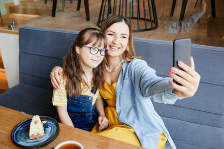 Candid portrait of happy mother and daughter with down syndrome taking selfie photo in cafe, copy spaceの写真素材